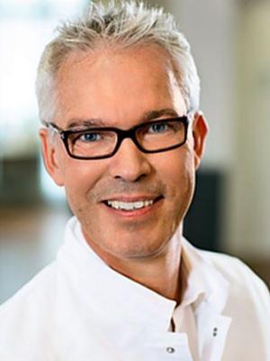 Porträt eines lächelnden, älteren Mannes mit grauen Haaren und schwarzer Brille, gekleidet in ein weißes Küchen- oder Hemd.